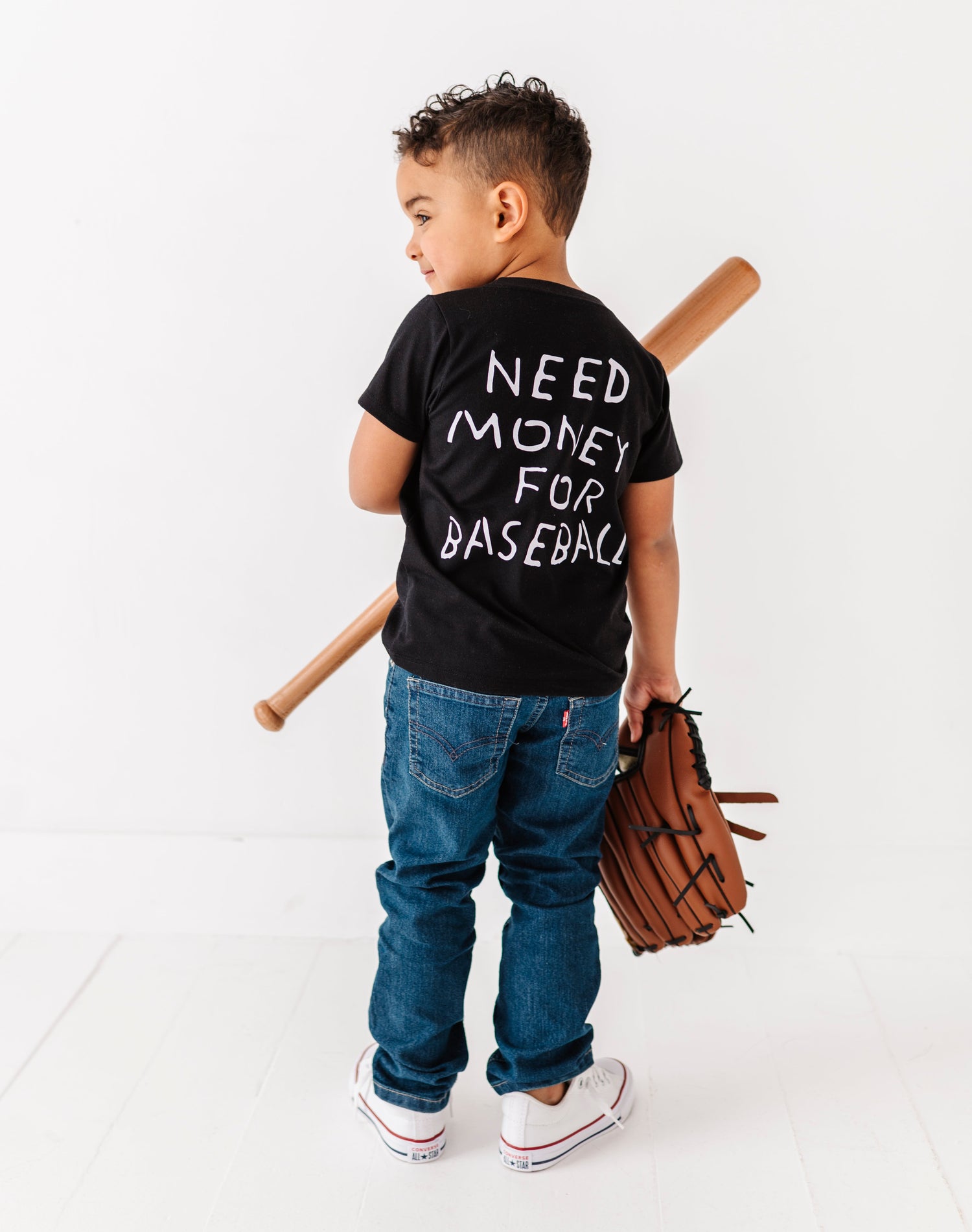 Child wearing a black t-shirt with 'Need Money for Baseball' text, holding baseball bats and a glove on a white background