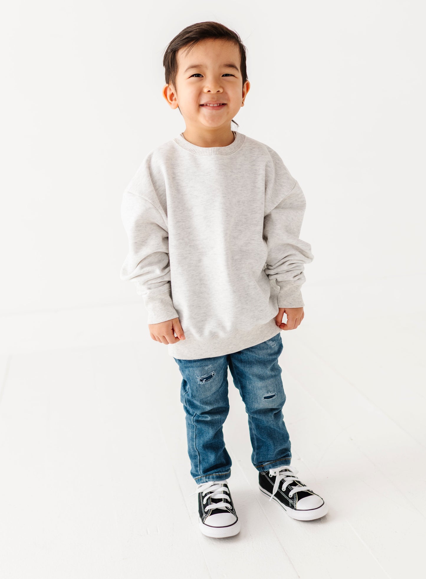 Child wearing a gray sweatshirt, blue jeans, and black and white sneakers on a white background